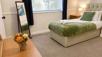 A neatly arranged bedroom with a bed, a bedside table, a lamp, a mirror, and a vase with flowers.