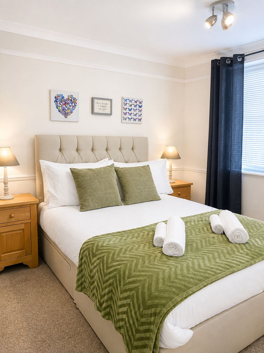 A neatly made bed with a green patterned bedspread, two pillows, and rolled towels on a wooden nightstand.
