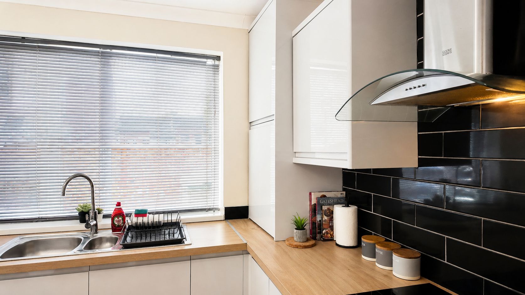 A modern kitchen with a stainless steel sink, a window with blinds, a range hood, and black tiled backsplash.