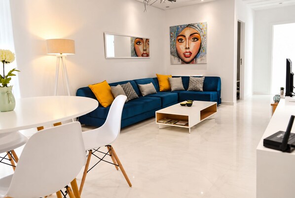 Ein modernes Wohnzimmer mit einem blauen Sofa, weißen Sesseln, einem Couchtisch und einem Gemälde an der Wand.