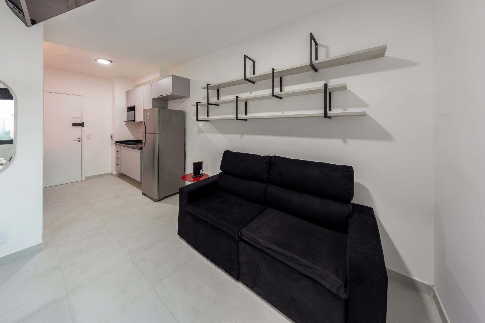 Ein stilvoller Wohnbereich mit einem schwarzen Sofa, einer Küche mit einem Edelstahlkühlschrank und einer weißen Wand mit schwarzen Regaleinheiten.
