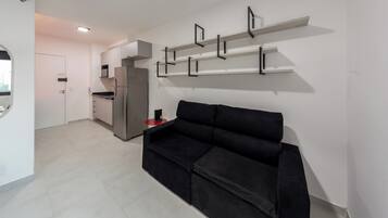 Ein stilvoller Wohnbereich mit einem schwarzen Sofa, einer Küche mit einem Edelstahlkühlschrank und einer weißen Wand mit schwarzen Regaleinheiten.