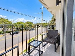 A balcony with a chair and a small table, overlooking a street with cars and trees. - Coastal Haven at 300 Tenth | 223 (Myrtle Beach)
