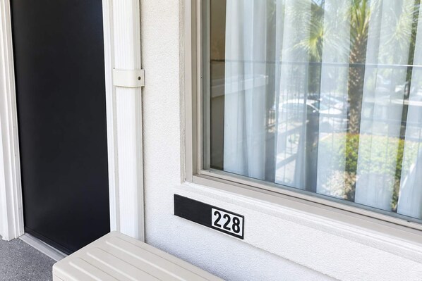 Uma porta com uma placa de identificação com o número 228 e uma janela que mostra palmeiras e uma cerca.