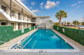 Una piscina con una profundidad de 3,5 pies, rodeada por un edificio de fachada blanca y un seto verde.