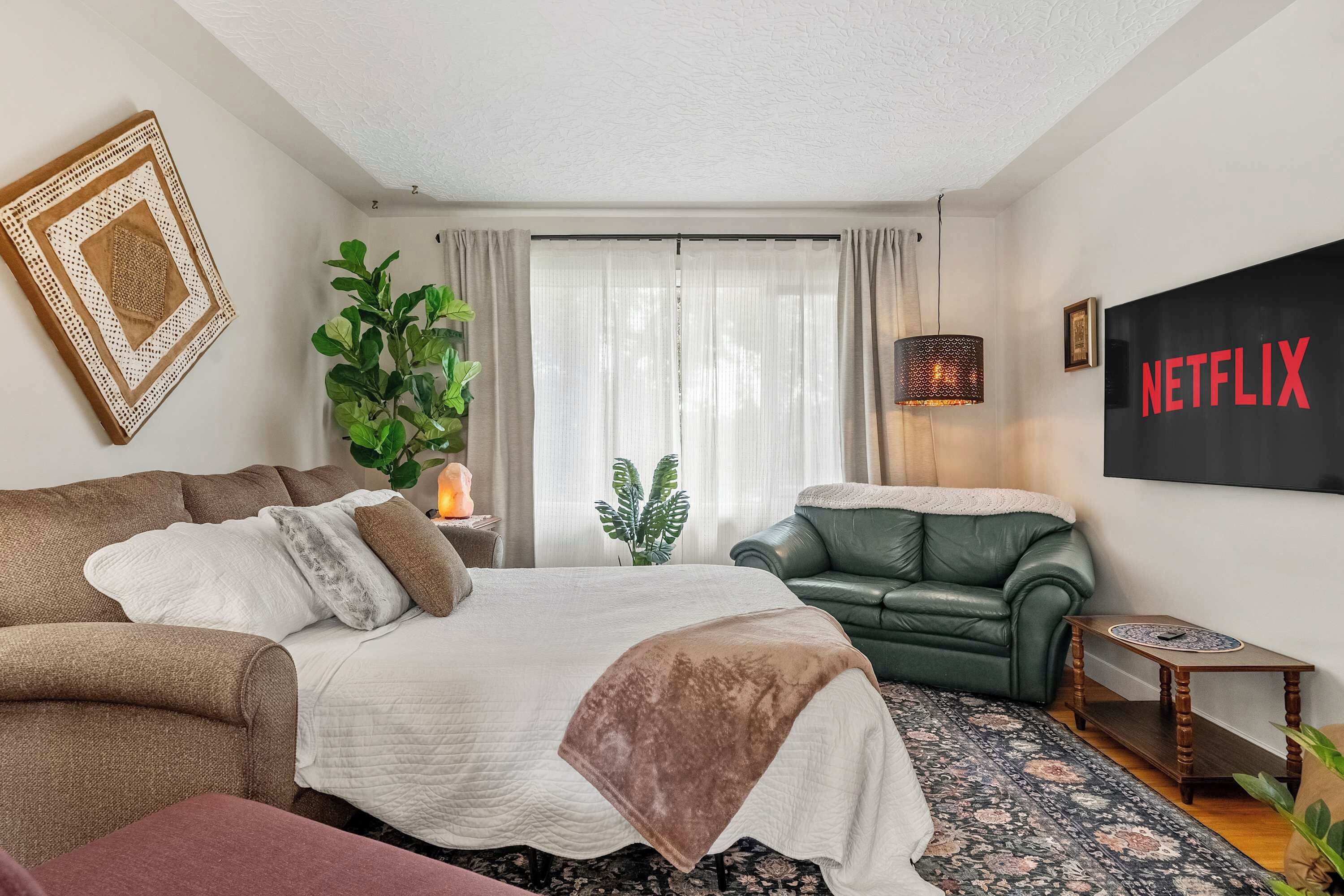 A bedroom with a bed, a green sofa, a wall-mounted TV, and a large window with curtains.