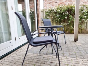 Eine Terrasse mit Tisch und zwei Stühlen, eine Glastür und eine Backsteinmauer mit blühenden Pflanzen.