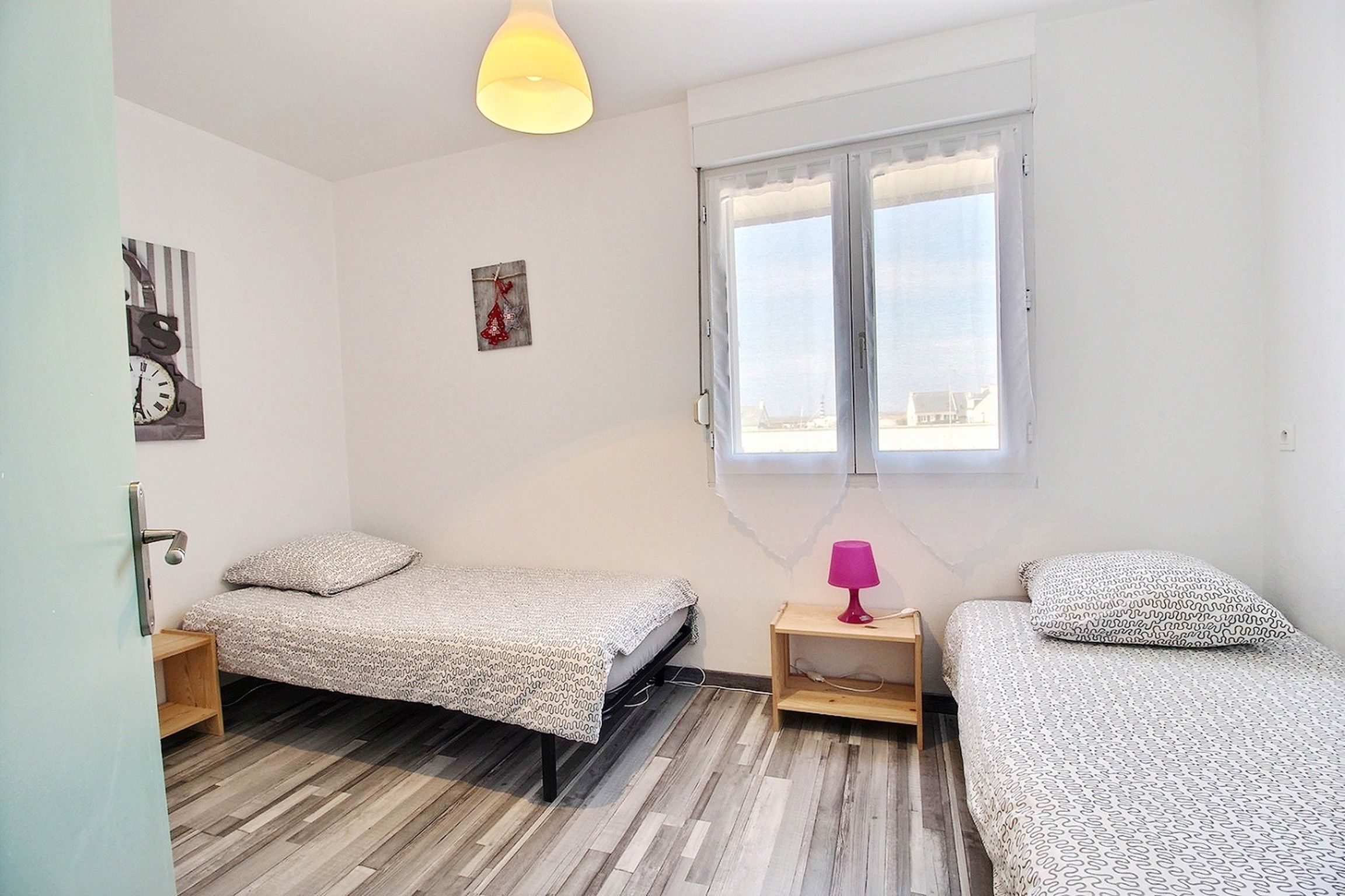 Une chambre avec deux lits simples, une petite table avec une lampe, une fenêtre avec des rideaux et une porte.