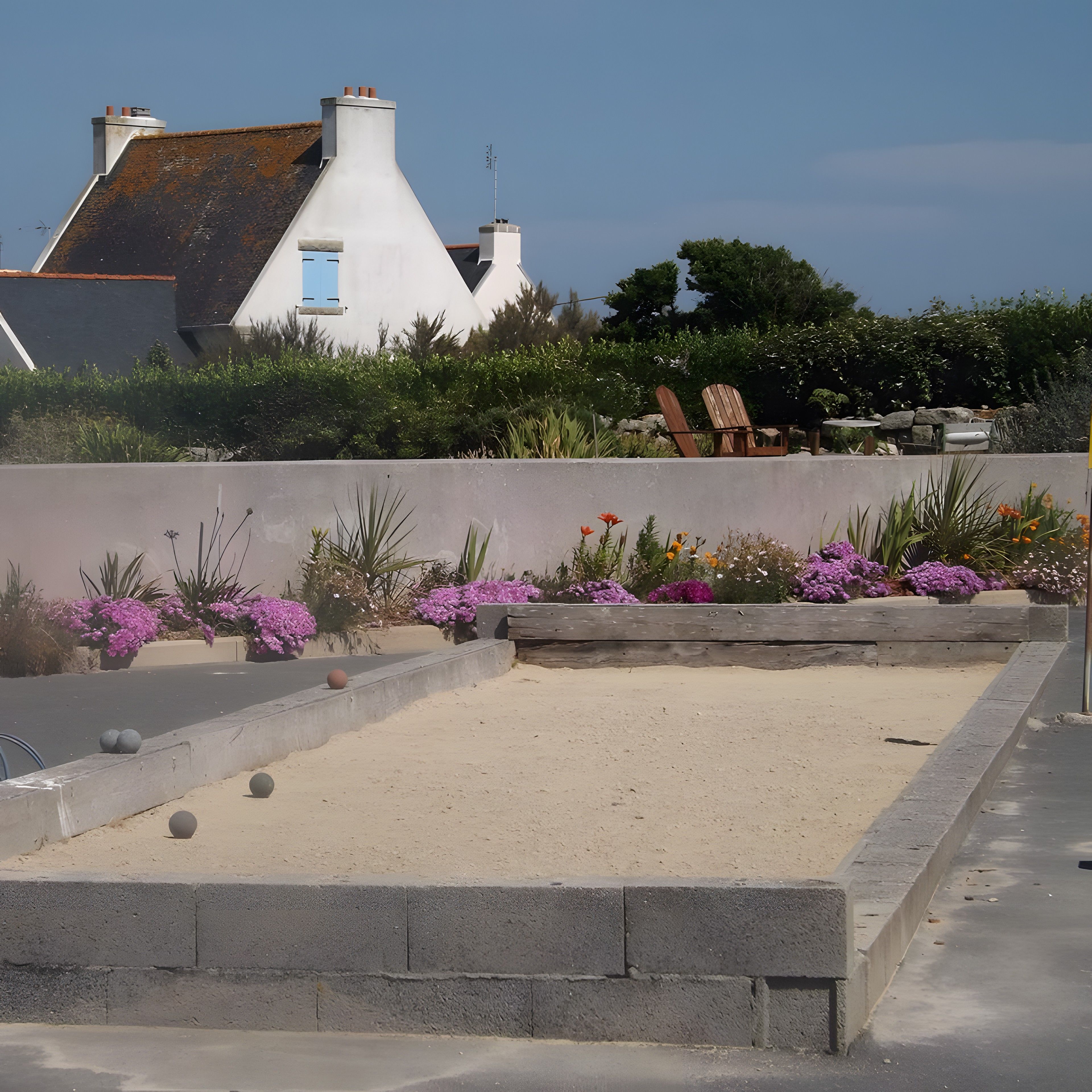 Une cour avec un bac à sable, des parterres de fleurs et une chaise en bois.