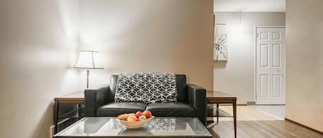 A living room with a glass coffee table, a black leather sofa with a patterned throw, a side table, a lamp, and a door in the background.