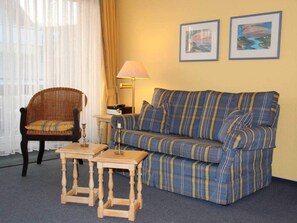 A room with a striped sofa, a wicker chair, a small wooden table, and two framed pictures on the wall. - Margeritenhaus Holiday Apartment 11 (Sylt)