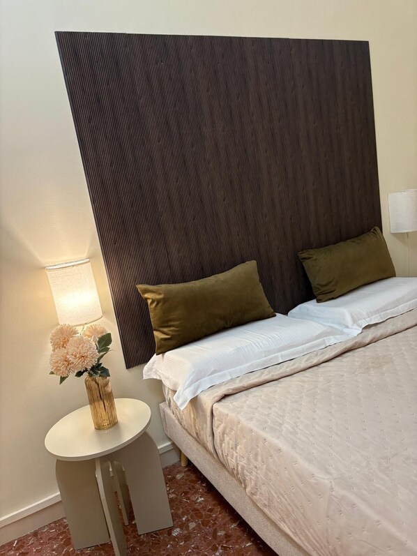 Una habitación de hotel con una cama, almohadas, una mesita de noche con una lámpara y un jarrón con flores.