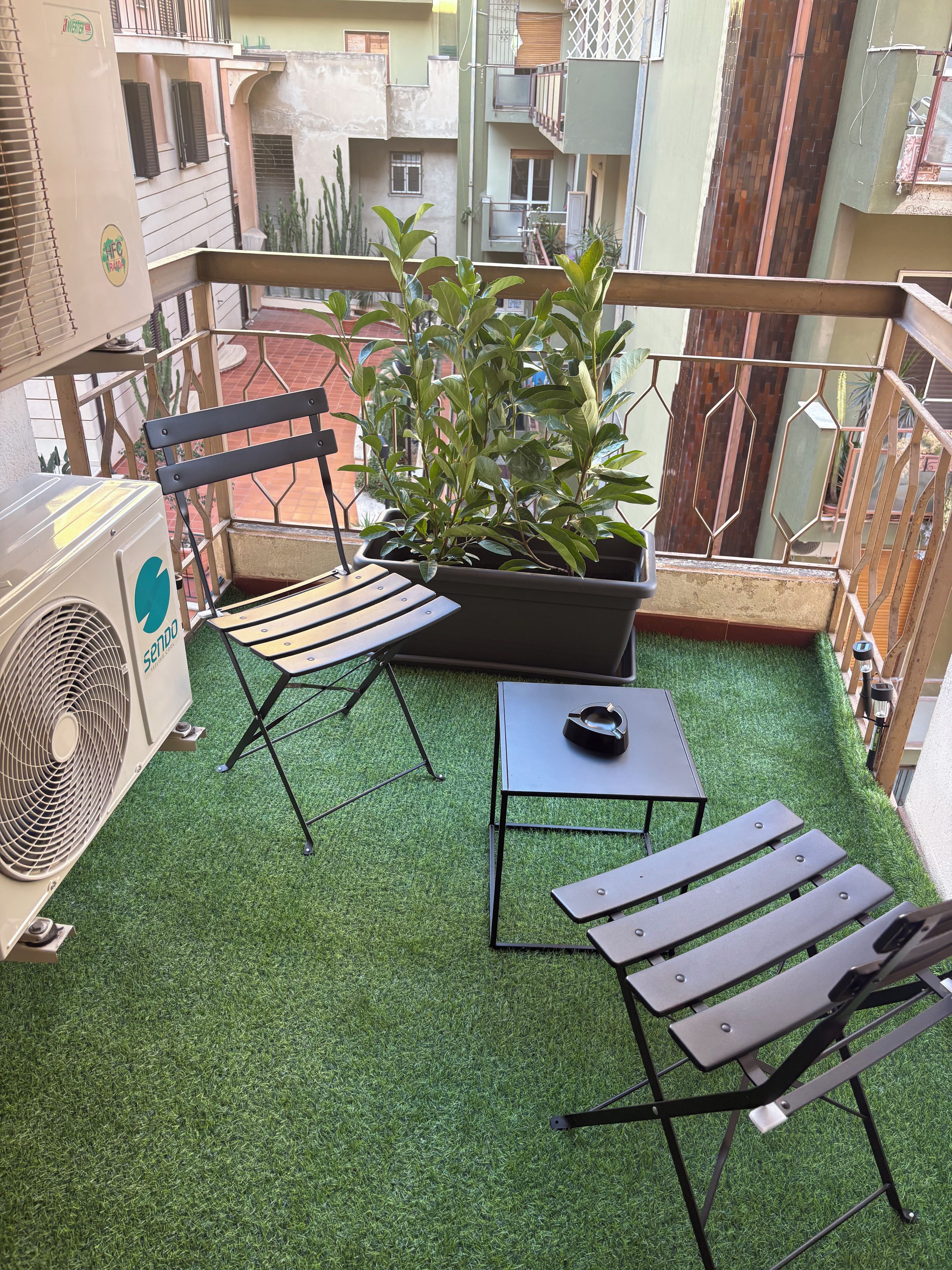 Un balcón con césped artificial, una mesa, dos sillas, una planta en maceta y un ventilador de pie.