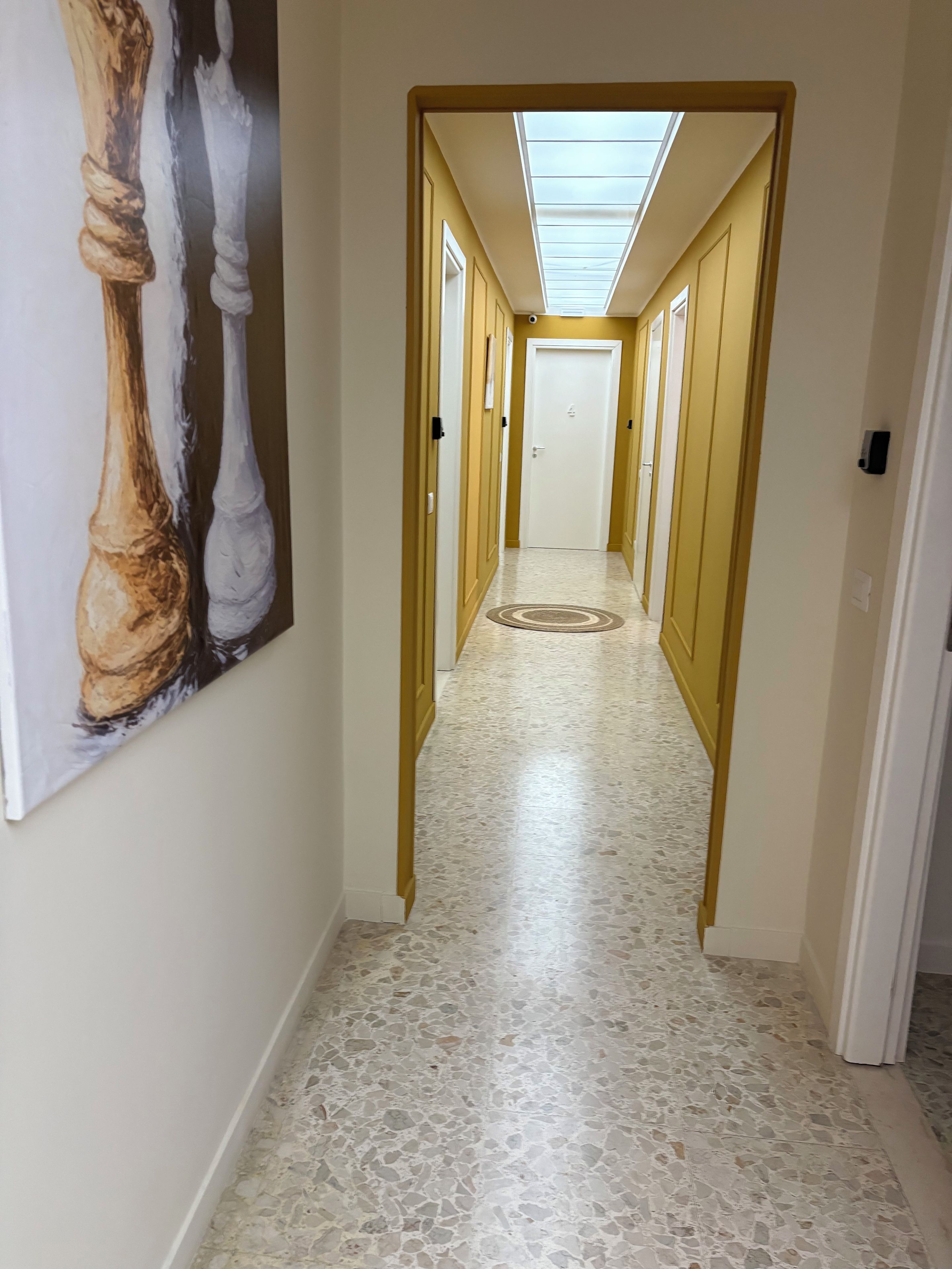 Un pasillo con piso de terrazo, un cuadro en la pared y un tragaluz arriba.