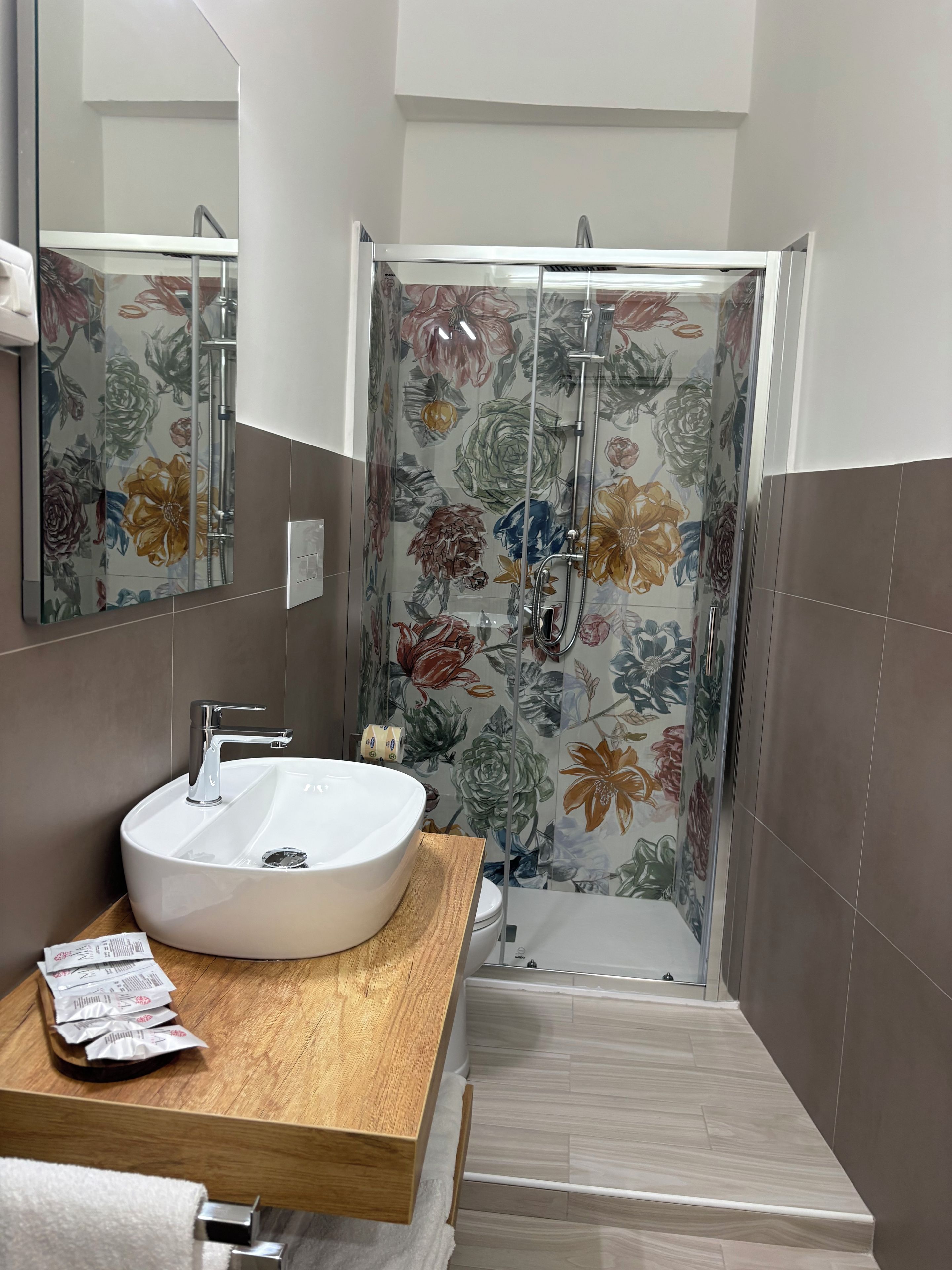 Un baño con un lavamanos grande y blanco, un mueble de tocador de madera, una ducha con puertas de vidrio con motivos florales y un espejo sobre el lavamanos.