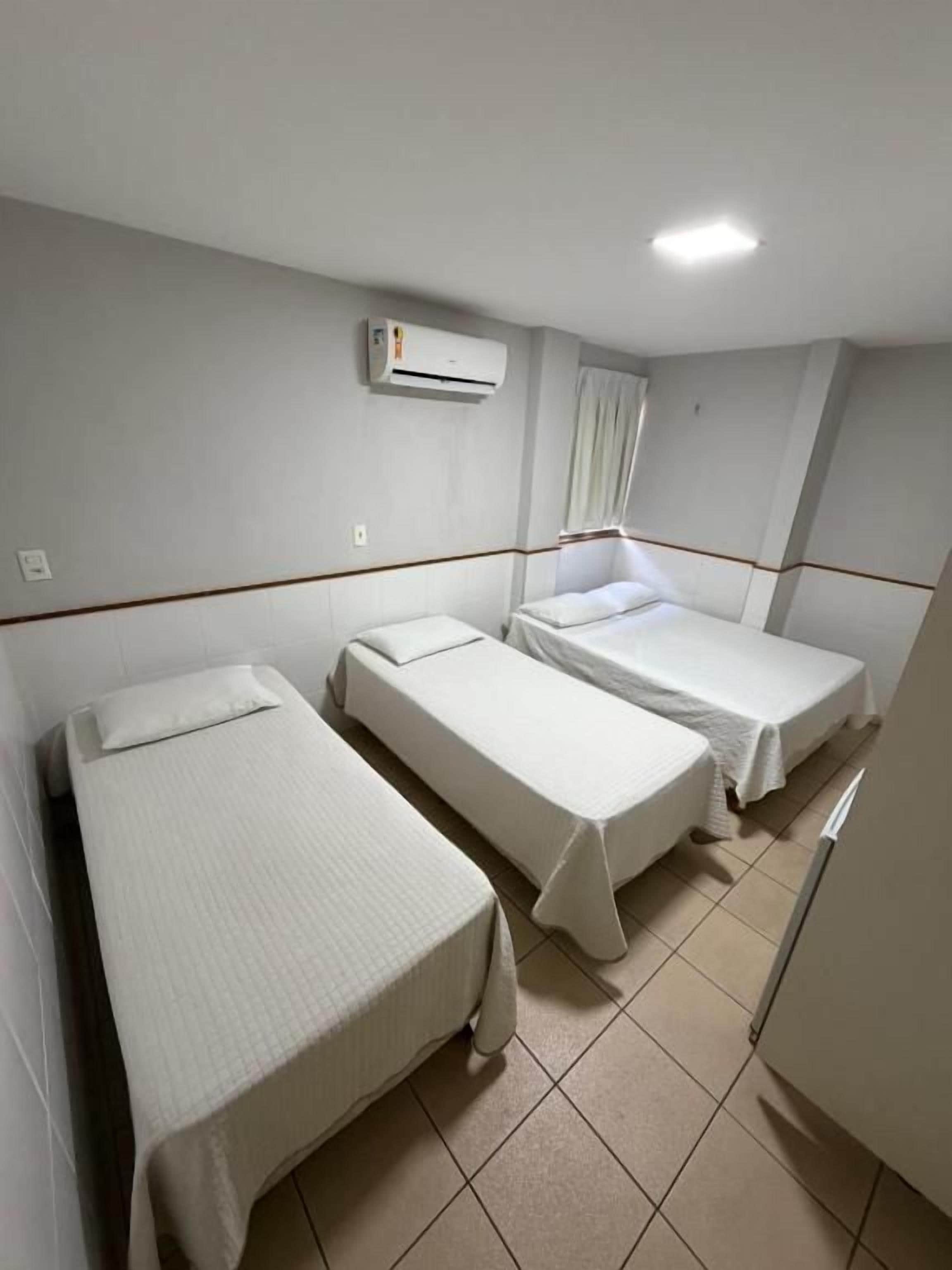 Habitación de hotel con tres camas individuales, ventilador de techo, unidad de aire acondicionado y ventana con cortinas.