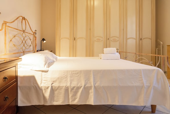 A neatly made bed with white linens, a wooden nightstand with a lamp, and a wardrobe with vertical stripes. - Emilia Suite Holiday (Modena)
