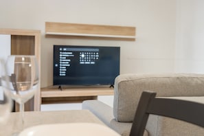 Una sala moderna con una televisión de pantalla plana montada en un estante de madera, una mesa de vidrio con una copa de vino y un sofá gris.