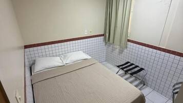 A small, single-bed room with a tiled floor, a window with curtains, and an air conditioning unit.