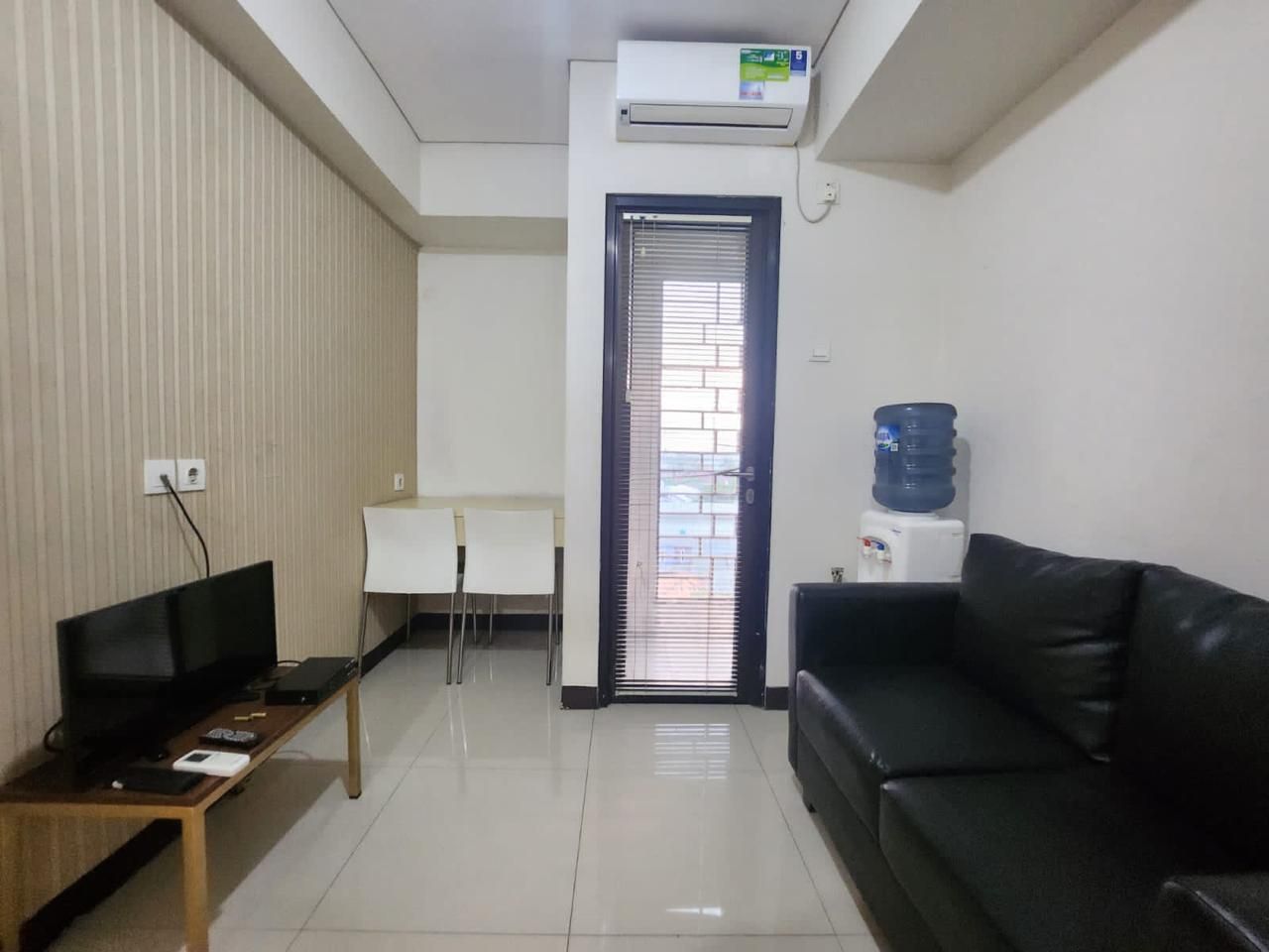 Habitación con televisión de pantalla plana, sofá de cuero negro, escritorio blanco y dispensador de agua.