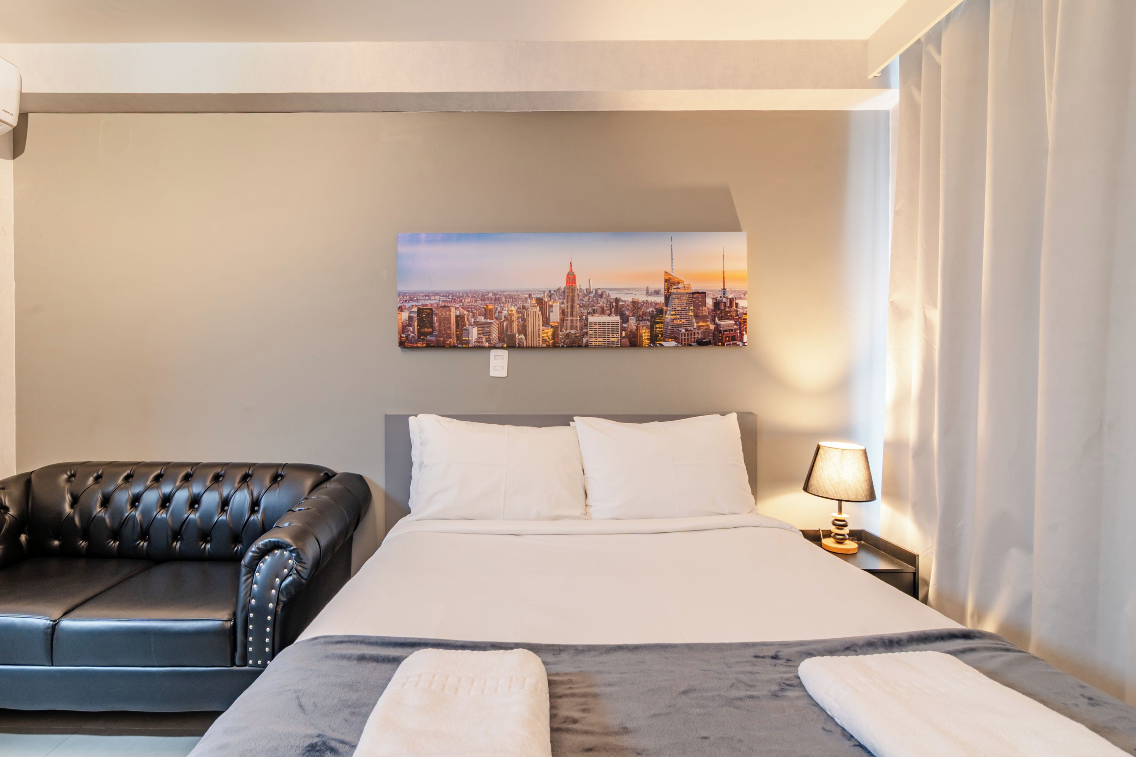 A hotel room with a bed, a black leather sofa, a cityscape painting on the wall, and a nightstand with a lamp.