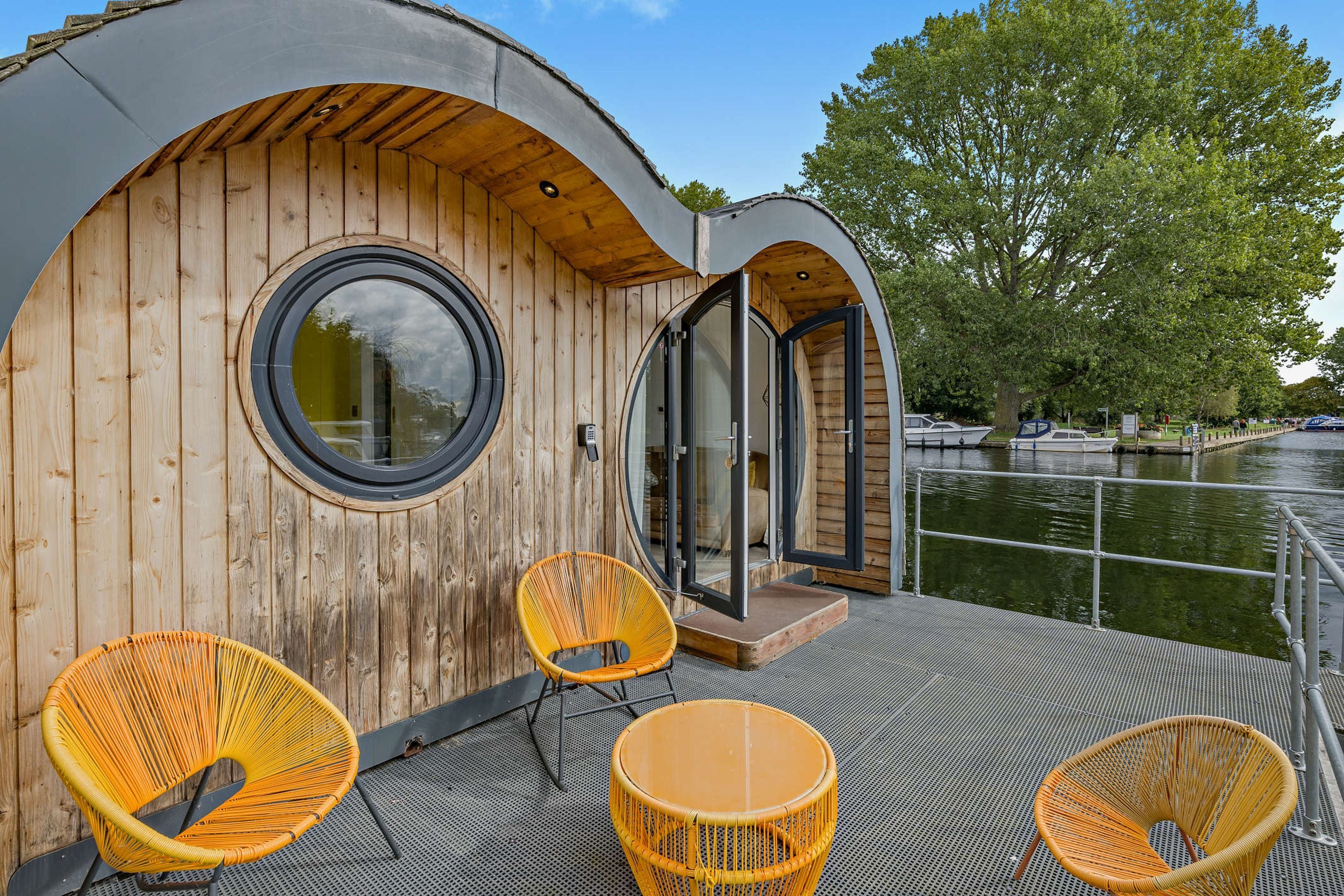 Un bote de madera con ventanal circular, una puerta y un pequeño espacio exterior con sillas de mimbre.