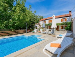 Ein Schwimmbecken mit Liegestühlen und Sonnenschirmen, umgeben von Grünpflanzen und einem Gebäude mit Ziegeldach.