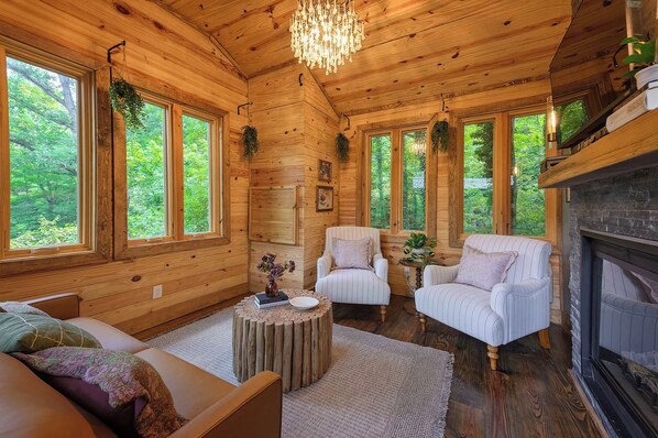 A cozy log cabin interior with a fireplace, two armchairs, a sofa, and large windows offering a view of the forest. - The Branch Luxury Treehouse with Private Sauna (Chattanooga)