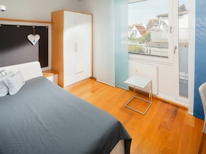 A bedroom with a bed, a wardrobe, a small table, and a window with a view of houses. - Holiday Apartment With 2 Bedrooms (Norderney)
