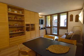 A dining area with a table, chairs, and a kitchen area with wooden cabinets and shelves. - Des Alpes 15 Studio Apartment MhD (Madonna di Campiglio)