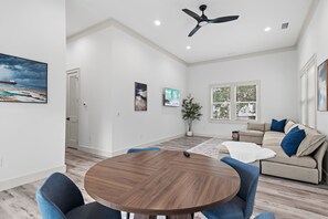 A spacious living room with a round wooden table, blue chairs, a beige sofa with blue cushions, and a ceiling fan. - 🐾The Lookout at Betty | Heated Pool & Covered Balconies & Pet Friendly & Bikes🚲 (Santa Rosa Beach)