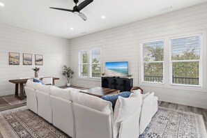 A modern living room with a sectional sofa, a coffee table, and a flat-screen TV. There are large windows with blinds, a ceiling fan, and artwork on the walls. - 🐾The Lookout at Betty | Heated Pool & Covered Balconies & Pet Friendly & Bikes🚲 (Santa Rosa Beach)