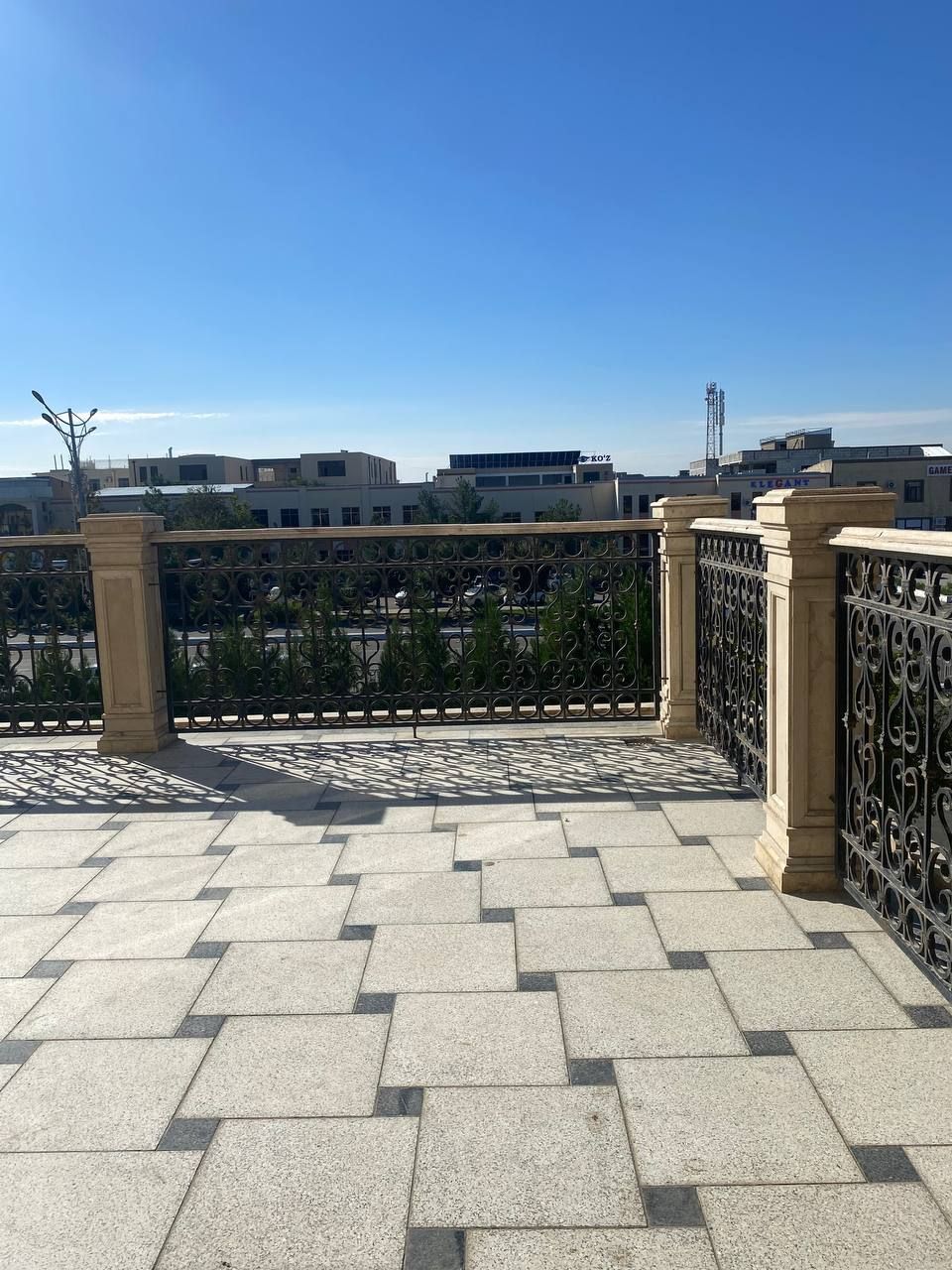 Una terraza empedrada con valla y pilares metálicos, con vistas a un paisaje urbano con edificios y cielo despejado.