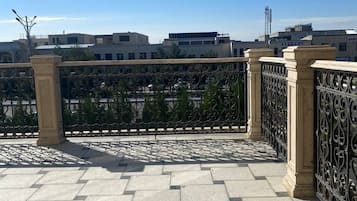 Una terraza empedrada con valla y pilares metálicos, con vistas a un paisaje urbano con edificios y cielo despejado.
