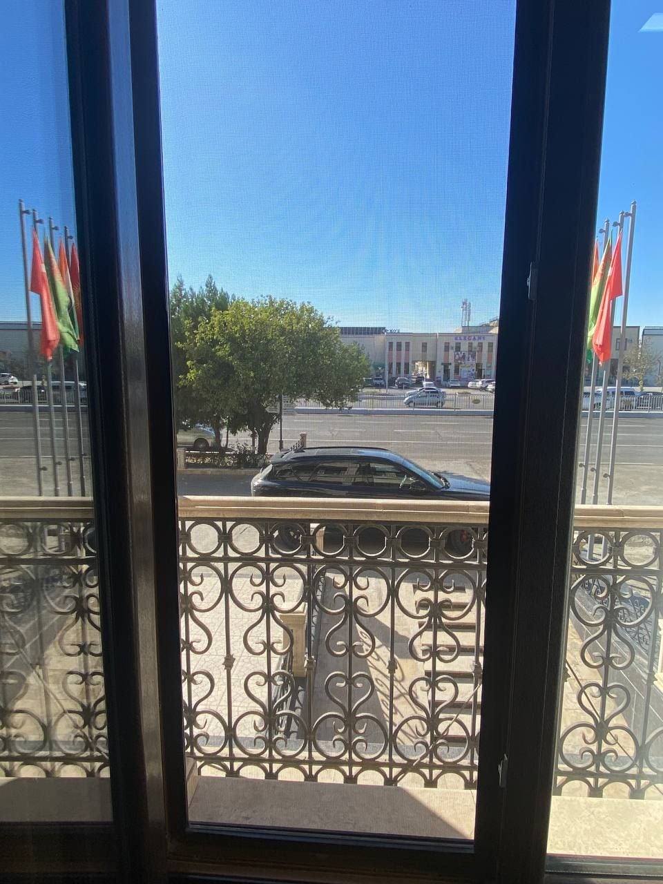 Vista a través de una ventana con barandilla de metal, un árbol y un edificio con banderas al fondo.