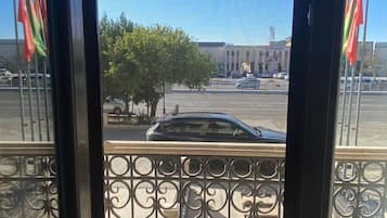 Vista a través de una ventana con barandilla de metal, un árbol y un edificio con banderas al fondo.