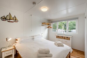 A bedroom with a bed, bedside tables, a hanging light fixture, a window with a view, and a wall-mounted shelf. - Host Stay Amazons (Beccles)