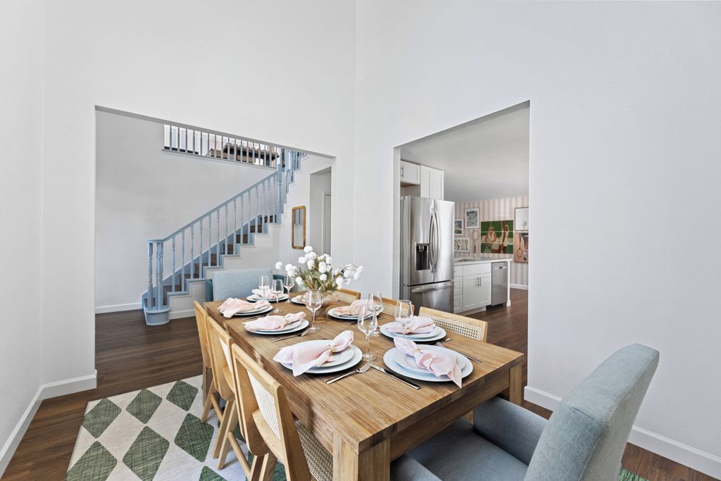 A dining area with a wooden table set for four, a staircase with blue railings, and a kitchen in the background.