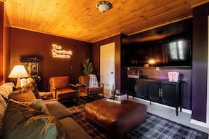 Ein gemütliches Wohnzimmer mit einem Ledersofa, einem karierten Teppich, einem Fernseher und einem Neon-Schild mit der Aufschrift 'The Overlook Theatre'.