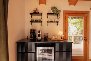 Ett köksutrymme med en kaffebryggare, muggar och en vattenkokare på bänkskivan. Ovanför bänkskivan finns tre hyllor, och på varje hylla står olika saker.