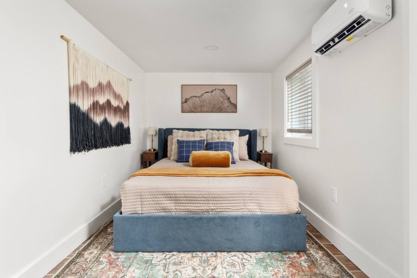 A bedroom with a bed, a wall-mounted air conditioner, a window with blinds, and a patterned rug.