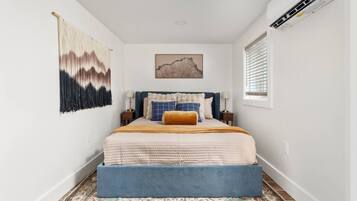 A bedroom with a bed, a wall-mounted air conditioner, a window with blinds, and a patterned rug.