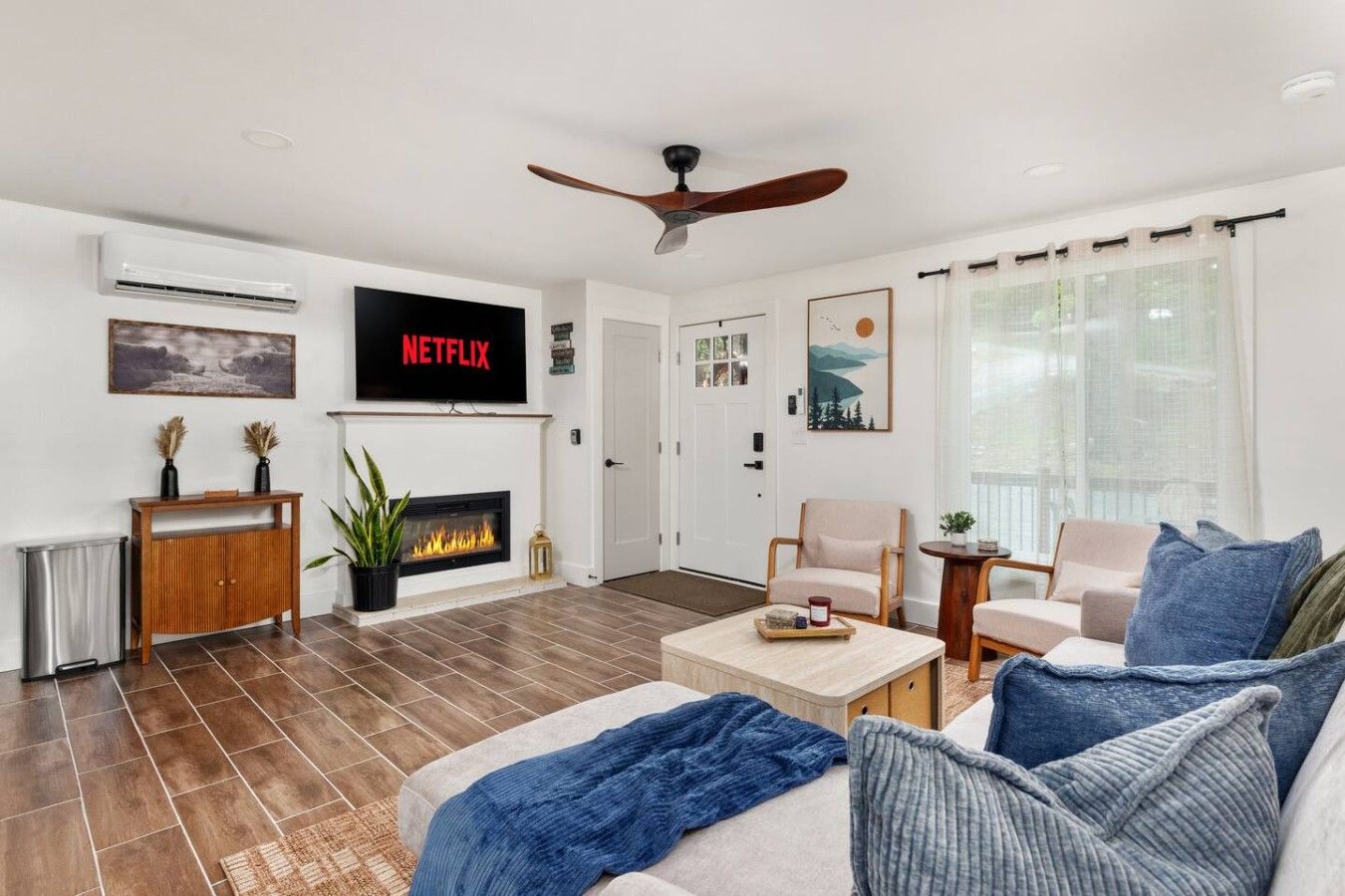 Un salon doté d’une cheminée, d’un ventilateur de plafond, d’un téléviseur fixé au mur et d’une grande fenêtre avec des rideaux.