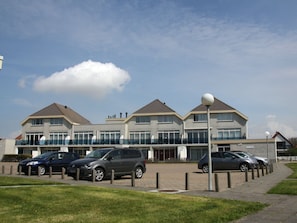 A modern building with multiple floors, a parking area with several cars, and a clear sky. - Seinpost 28 Callantsoog (Callantsoog)
