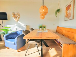 A room with a wooden table, blue armchair, and a bench. There are potted plants and a wall-mounted light fixture. - Seinpost 28 Callantsoog (Callantsoog)