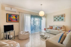 A living room with a flat-screen TV, a bean bag chair, a sofa with colorful cushions, a glass coffee table, and a wall with a framed artwork. - Apartment 'Areias Do Mar' (Albufeira)