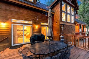 A wooden deck with a table and chairs, a covered grill, and a sliding glass door leading to an interior space with a sofa and a coffee table. - Charming 4BR Tahoe Cabin Near Speedboat Beach, Kings Beach & Skiing (Kings Beach)