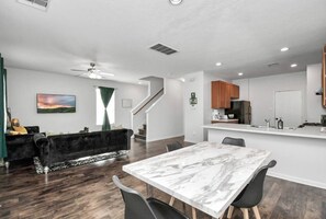 A modern living room with a dark sofa, a dining table with chairs, a kitchen area with wooden cabinets, and a staircase leading to another level. - Beeline Living (Houston)