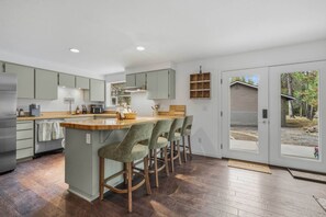 A modern kitchen with a central island, bar stools, and a sliding glass door leading to a covered outdoor area. - Cozy River Retreat on Deschutes: Hot Tub, Dog Friendly, Game Room + Quality Beds (Three Rivers)