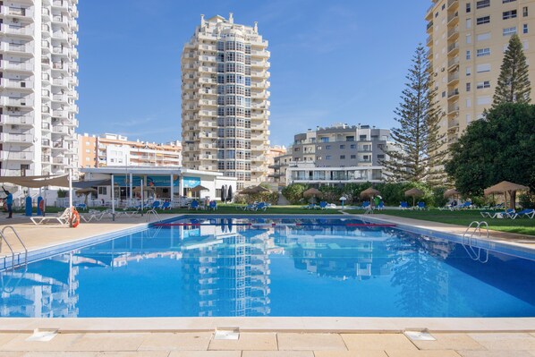 Una piscina amb aigua transparent de color blau, envoltada de gandules i ombrel·les, amb edificis alts al fons.